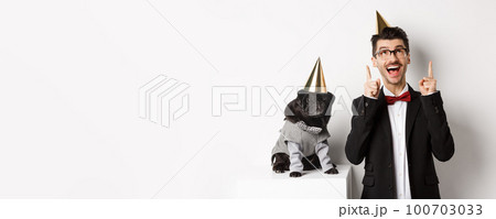 Cute black pug dog wearing party cone and standing near happy owner, man pointing fingers up at copy space, celebrating birthday, white background Cute black pug dog wearing party cone and standing near happy owner, man pointing fingers up at copy space, celebrating birthday, white background 100703033