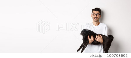 Excited hipster guy with his dog looking left at logo. Handsome pet owner carry black pug and staring at promo banner, standing over white background 100703602