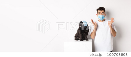 Covid-19, animals and quarantine concept. Shocked dog owner and pug wearing medical masks, staring at camera amazed, standing over white background 100703603