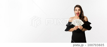 Beauty and shopping concept. Excited woman in black dress, showing money prize and staring happy at camera, standing over white background Beauty and shopping concept. Excited woman in black dress, showing money prize and staring happy at camera, standing over white background 100703605