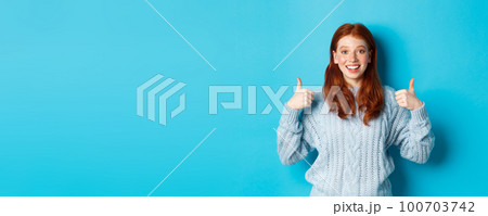 Cheerful teen girl with red hair, showing thumbs up in approval, like and praise gesture, standing over blue background 100703742