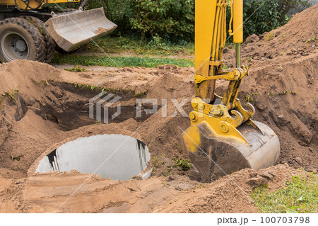 A bucket of excavator with a pile of sand and earth buries sewer concrete rings in the industrial zone A bucket of excavator with a pile of sand and earth buries sewer concrete rings in the industrial zone 100703798