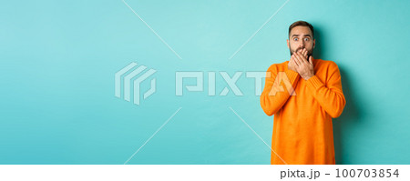 Startled man looking at horrible scene, gasping and stare scared, standing in orange winter sweater, studio background 100703854