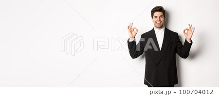 Concept of new year party, celebration and lifestyle. Portrait of successful handsome businessman in suit, winking and showing okay signs, recommending something good, white background 100704012
