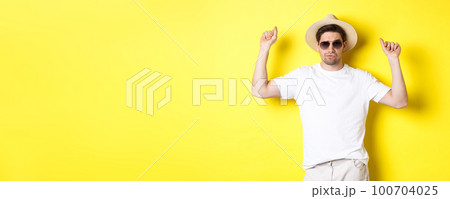 Tourism, travelling and holidays concept. Man tourist enjoying vacation, dancing in straw hat and sunglasses, posing against yellow background 100704025