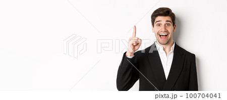 Close-up of handsome businessman in black suit, smiling amazed, showing number one, standing over white background 100704041