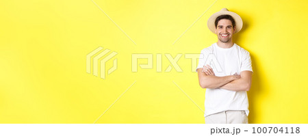 Tourism. Handsome young man looking happy, wearing straw hat for travelling, cross hands on chest and smiling, standing over yellow background Tourism. Handsome young man looking happy, wearing straw hat for travelling, cross hands on chest and smiling, standing over yellow background 100704118
