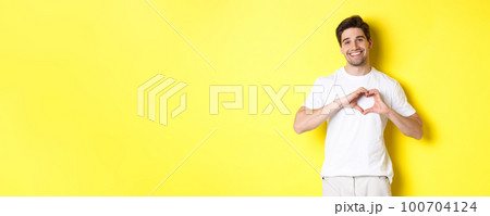 Happy romantic man showing heart sign, smiling and express love, standing over yellow background 100704124