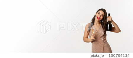 Winter holidays celebration concept. Happy woman on Christmas party saying cheers, raising bottle of champagne and smiling, standing in elegant dress, white background 100704134