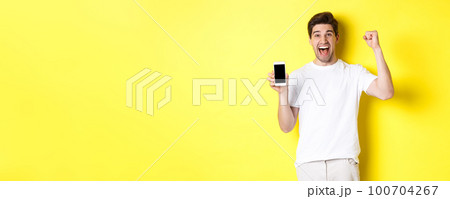 Cheerful guy showing smartphone screen, raising hand up and celebrating, triumphing over internet achievement, standing over yellow background Cheerful guy showing smartphone screen, raising hand up and celebrating, triumphing over internet achievement, standing over yellow background 100704267