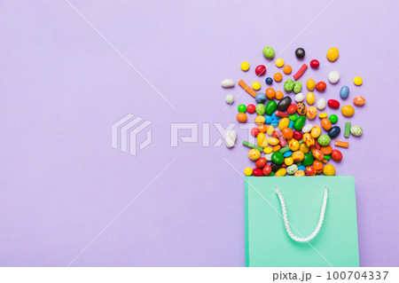 shopping paper gift bag in corner full of assorted traditional candies falling out on colored background with copy space. Happy Holidays sale concept shopping paper gift bag in corner full of assorted traditional candies falling out on colored background with copy space. Happy Holidays sale concept 100704337