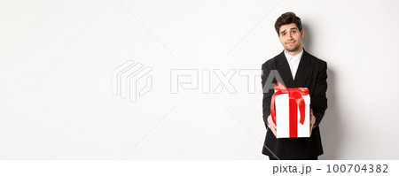Concept of christmas holidays, celebration and lifestyle. Image of handsome boyfriend in black suit, giving you a present, extending hands with gift, standing over white background 100704382