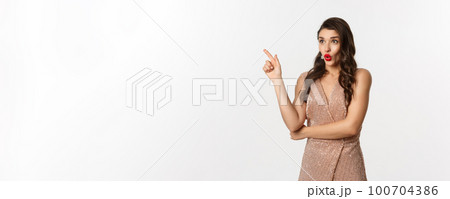 Christmas, holidays and celebration concept. Surprised girlfriend in evening dress, saying wow and pointing finger at promo offer, starring at logo, standing over white background 100704386