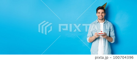 Smiling man holding b-day cake and wearing birthday party hat, celebrating over blue background 100704396