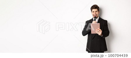 Image of handsome man in black suit, singing karaoke on digital tablet, holding microphone, standing over white background 100704439