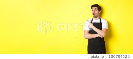 Waiter in black apron checking out promo offer, pointing finger and looking left at your logo, standing over yellow background 100704529