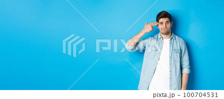 Tired and annoyed adult man making finger gun gesture near head, shooting himself from boredom, standing against blue background Tired and annoyed adult man making finger gun gesture near head, shooting himself from boredom, standing against blue background 100704531