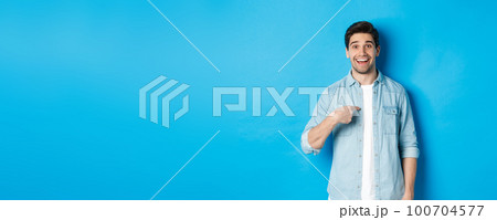 Happy and surprised man pointing at himself, smiling pleased, standing against blue background Happy and surprised man pointing at himself, smiling pleased, standing against blue background 100704577
