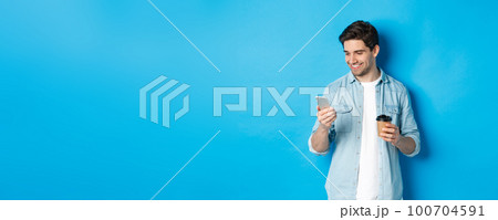 Handsome modern guy reading message on cell phone and drinking coffee, standing against blue background 100704591