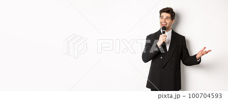 Portrait of handsome man in black suit singing a song, holding microphone and giving speech, standing against white background 100704593