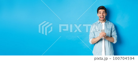 Surprised and happy man winning something online, holding smartphone and rejoicing, standing against blue background 100704594