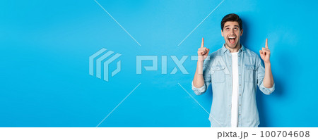 Excited handsome man pointing and looking up, showing advertisement, standing over blue background Excited handsome man pointing and looking up, showing advertisement, standing over blue background 100704608