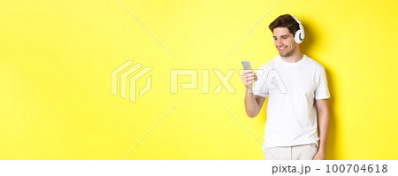 Handsome modern guy choosing playlist on smartphone, wearing headphones, standing over yellow background Handsome modern guy choosing playlist on smartphone, wearing headphones, standing over yellow background 100704618