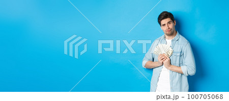 Handsome young man keeping money to himself, smiling and looking greedy, standing over blue background 100705068