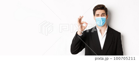 Concept of covid-19, business and social distancing. Close-up of good-looking businessman in trendy suit and medical mask, showing okay sign and winking, standing against white background Concept of covid-19, business and social distancing. Close-up of good-looking businessman in trendy suit and medical mask, showing okay sign and winking, standing against white background 100705211