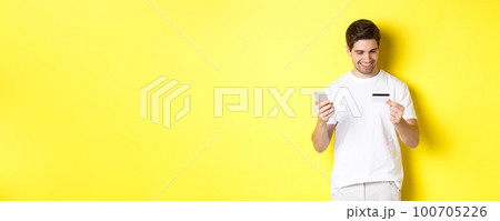 Guy making online order, register credit card in mobile application, holding smartphone and smiling, standing over yellow background Guy making online order, register credit card in mobile application, holding smartphone and smiling, standing over yellow background 100705226
