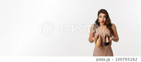 Party and celebration concept. Beautiful woman in elegant dress asking help, showing two different heels, clueless what to wear, standing over white background 100705262