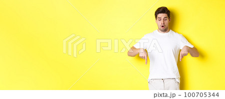 Worried guy pointing fingers and looking down, standing startled against yellow background 100705344