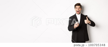 Portrait of handsome man in trendy suit showing thumbs-up in approval, using mobile phone app, smiling pleased, standing over white background 100705348