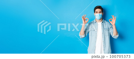 Concept of covid-19, pandemic and social distancing. Satisfied man in medical mask, showing ok signs in approval, agree or like something good, standing over blue background 100705373