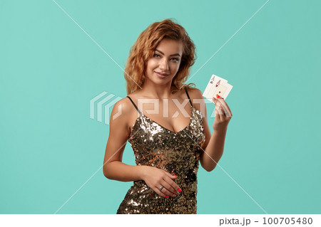 Ginger girl in shiny golden dress is posing holding two playing cards in her hands standing against a blue studio background. Casino, poker. Close-up. Ginger girl in shiny golden dress is posing holding two playing cards in her hands standing against a blue studio background. Casino, poker. Close-up. 100705480