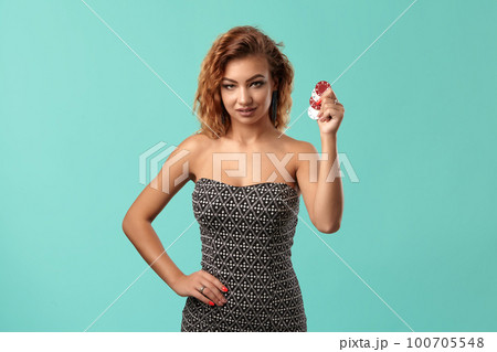 Ginger girl wearing gray dress is posing holding some chips in her hands standing against a blue studio background. Casino, poker. Close-up. 100705548