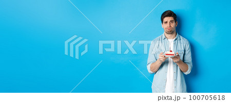 Upset and gloomy man holding birthday cake, sulking sad, standing against blue background 100705618