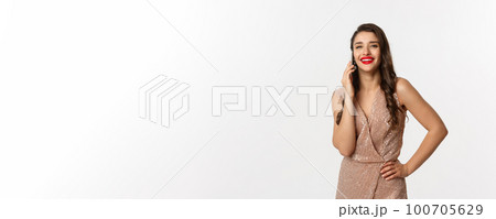 Christmas party and celebration concept. Attractive young woman with red lips, wearing luxury dress, talking on mobile phone and smiling, standing over white background Christmas party and celebration concept. Attractive young woman with red lips, wearing luxury dress, talking on mobile phone and smiling, standing over white background 100705629