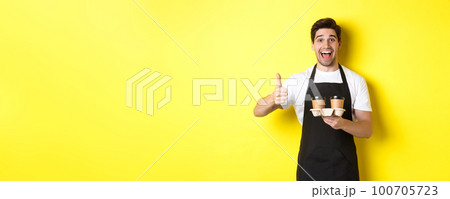 Cheerful barista recommend his cafe, holding takeaway coffee cups and showing thumbs up, standing against yellow background Cheerful barista recommend his cafe, holding takeaway coffee cups and showing thumbs up, standing against yellow background 100705723