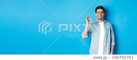 Attractive young man wearing glasses and casual clothes, showing ok good sign in approval, like something, standing against blue background 100705761