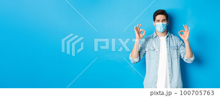 Concept of coronavirus, quarantine and social distancing. Cheeky man in medical mask winking, showing okay signs, assure or guarantee something, like and approve Concept of coronavirus, quarantine and social distancing. Cheeky man in medical mask winking, showing okay signs, assure or guarantee something, like and approve 100705763