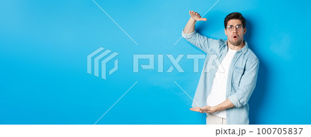 Confused handsome man showing big size, shaping large box and looking amazed, standing over blue background Confused handsome man showing big size, shaping large box and looking amazed, standing over blue background 100705837