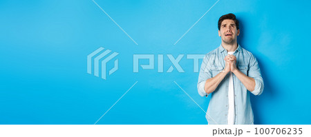 Desperate man begging and crying for help, pleading to god, looking up and holding hands in pray, standing against blue background 100706235
