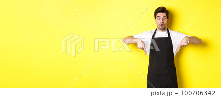 Surprised male barista in black apron checking out promo offer, pointing fingers down at logo banner, standing over yellow background Surprised male barista in black apron checking out promo offer, pointing fingers down at logo banner, standing over yellow background 100706342