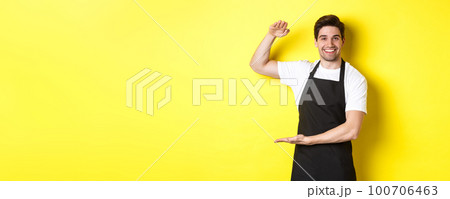 Smiling handsome barista showing something long or large, standing over yellow background 100706463