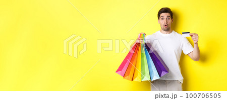 Excited man shopping on black friday, holding paper bags and credit card, standing against yellow background Excited man shopping on black friday, holding paper bags and credit card, standing against yellow background 100706505