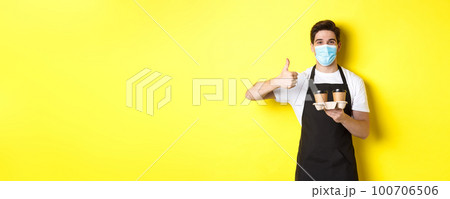 Concept of covid-19, cafe and social distancing. Young male barista in medical mask and black apron holding takeaway coffee cups, showing thumb up, yellow background 100706506