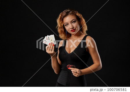 Ginger girl in a dark dress posing holding playing cards and chips in her hands standing against a black studio background. Casino, poker. Close-up. Ginger girl in a dark dress posing holding playing cards and chips in her hands standing against a black studio background. Casino, poker. Close-up. 100706581