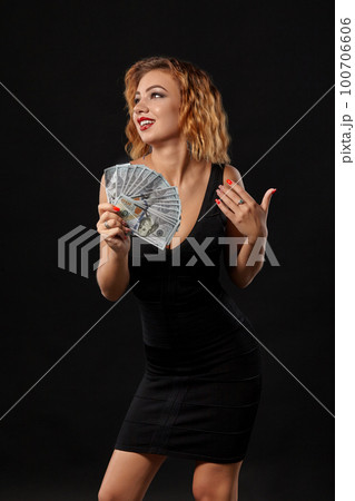 Ginger girl in dark dress posing holding fan of hundred-dollar bills in her hands standing against a black studio background. Casino, poker. Close-up. 100706606