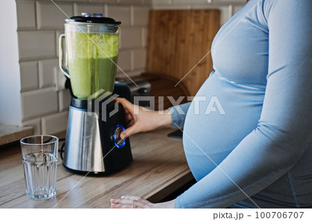 Green Smoothies Recipes For Pregnancy and Postpartum, Prenatal Nutrition. Pregnant woman preparing green vitamin smoothie with a blender and drinking in the kitchen. 100706707